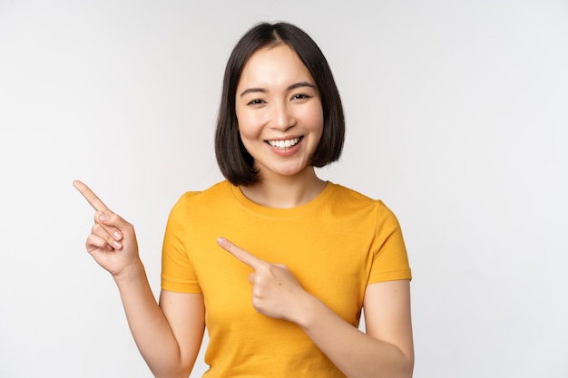 Портрет улыбающейся азиатской брюнетки в желтой футболке, указывающей пальцем влево, показывая промо-сделку с копией пространства, демонстрируя баннер, стоящий на белом фоне