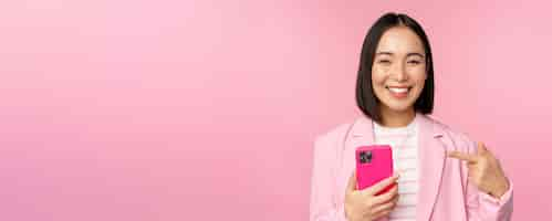 Free photo portrait of smiling asian businesswoman pointing at her mobile phone recommending smartphone app application on cellphone standing over pink background