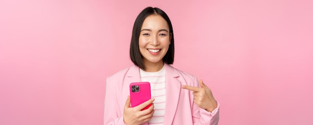 분홍색 배경 위에 서 있는 휴대폰에 스마트폰 앱 응용 프로그램을 추천하는 휴대폰을 가리키며 웃고 있는 아시아 여성 사업가의 초상화