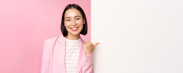 Free photo portrait of smiling asian businesswoman in suit corporate lady pointing finger at white empty wall b