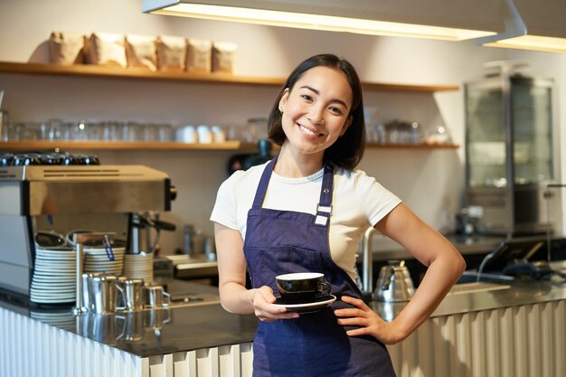 一杯のお茶を持ってコーヒーを作り、それをカフェ・クリに持っていく笑顔のアジアの女性バリスタのポートレート