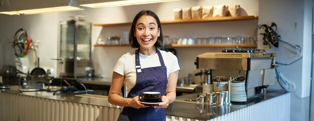 Portrait of smiling asian female barista making coffee holding cup of tea and taking it to cafe