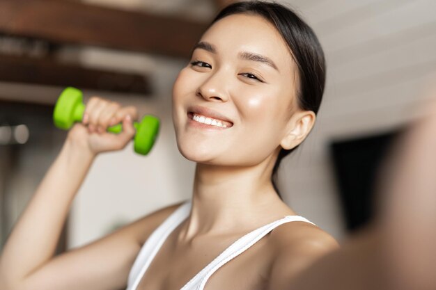 Portrait of smiling asian fitness instructor taking selfie with dumbbell athletic woman making photo...