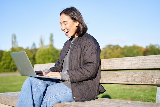 Free photo portrait of smiling asian girl sits on bench in park talks to friend online via laptop video chats u