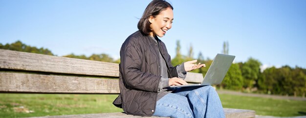 Free photo portrait of smiling asian girl sits on bench in park talks to friend online via laptop video chats
