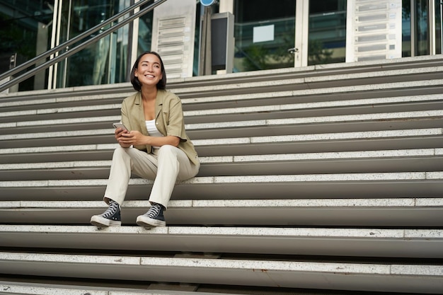 Free photo portrait of smiling asian girl sits on stairs outdoors sending message using smartphone app looking