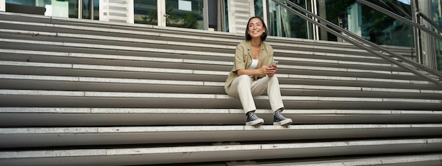 Free photo portrait of smiling asian girl sits on stairs outdoors sending message using smartphone app looking