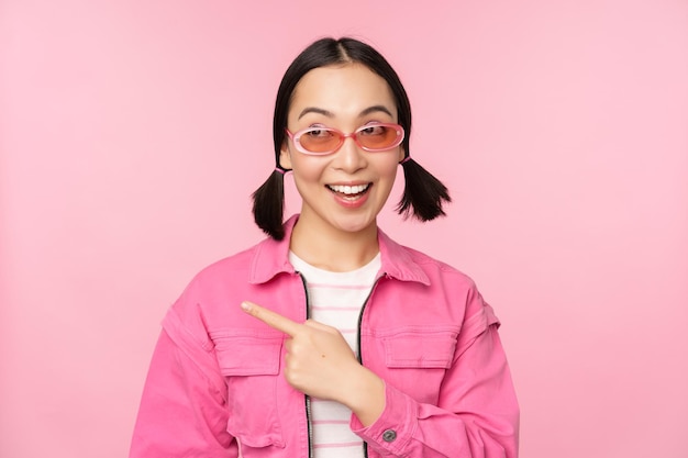 ピンクの背景の上に立っている広告バナーを示す左指を指しているスタイリッシュな衣装のサングラスで笑顔のアジアの女の子の肖像画コピースペース