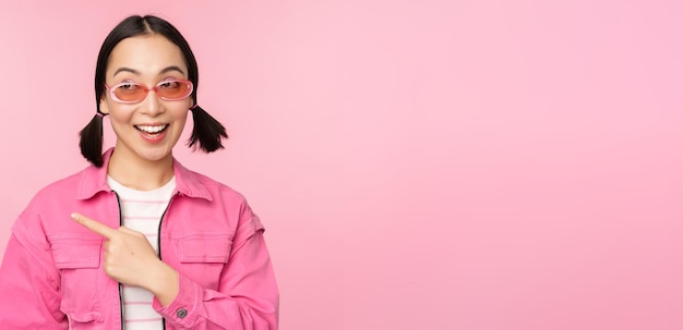 Portrait of smiling asian girl in stylish outfit sunglasses pointing finger left showing advertisement banner standing over pink background Copy space