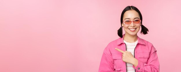 분홍색 배경 복사 공간 위에 서 있는 광고 배너를 보여주는 왼쪽 손가락을 가리키는 세련된 복장 선글라스를 쓴 웃는 아시아 소녀의 초상화