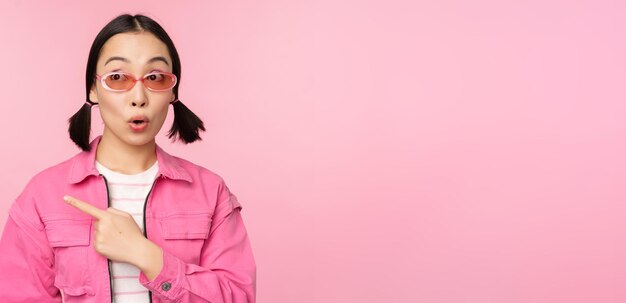 분홍색 배경 위에 서 있는 광고 배너를 보여주는 왼쪽 손가락을 가리키는 세련된 복장 선글라스를 끼고 웃고 있는 아시아 소녀의 초상화