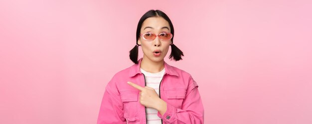 Portrait of smiling asian girl in stylish outfit sunglasses pointing finger left showing advertisement banner standing over pink background