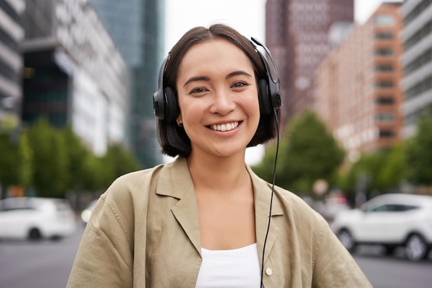 市内中心部の通りに立っているヘッドフォンで笑顔のアジアの女性の肖像画は、幸せそうに見えて耳を傾けます