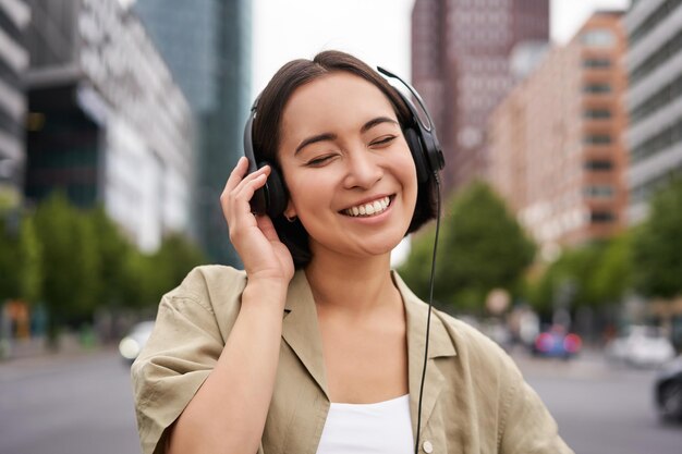市内中心部の通りに立っているヘッドフォンで笑顔のアジアの女性の肖像画は、幸せそうに見えて耳を傾けます