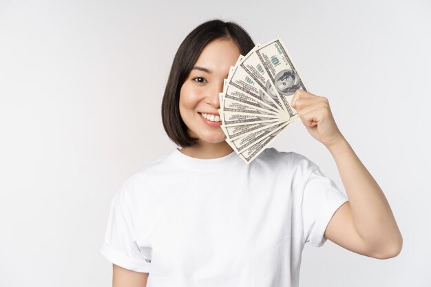 Portrait of smiling asian woman holding dollars money concept of microcredit finance and cash standing over white background