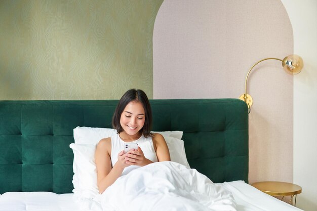 Портрет улыбающейся азиатской женщины, лежащей в постели и смотрящей на смартфон с помощью мобильного телефона со счастливым лицом