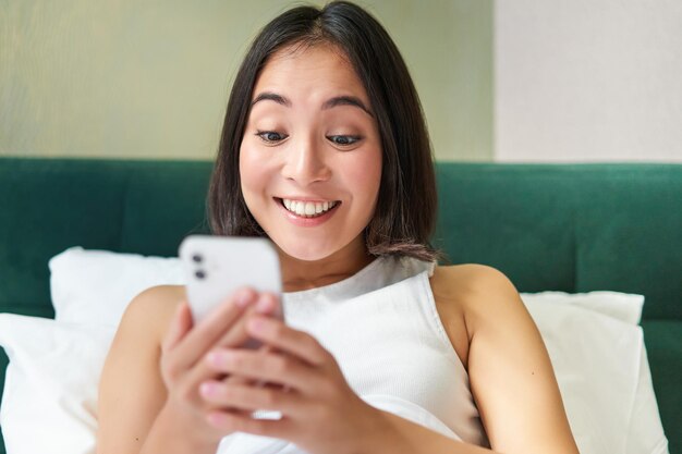 Portrait of smiling asian woman lying in bed looking at smartphone using mobile phone with happy fac