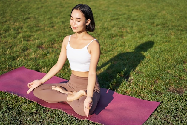 Free photo portrait of smiling asian woman meditating doing yoga on fresh air relaxing on rubber mat exercising