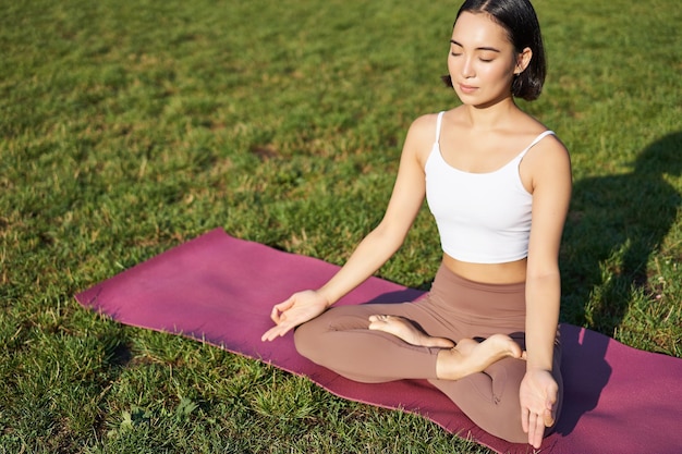 Free photo portrait of smiling asian woman meditating doing yoga on fresh air relaxing on rubber mat exercising