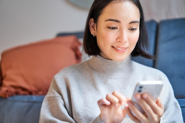 Free photo portrait of smiling asian woman sits at home uses smartphone app orders delivery messages someone on