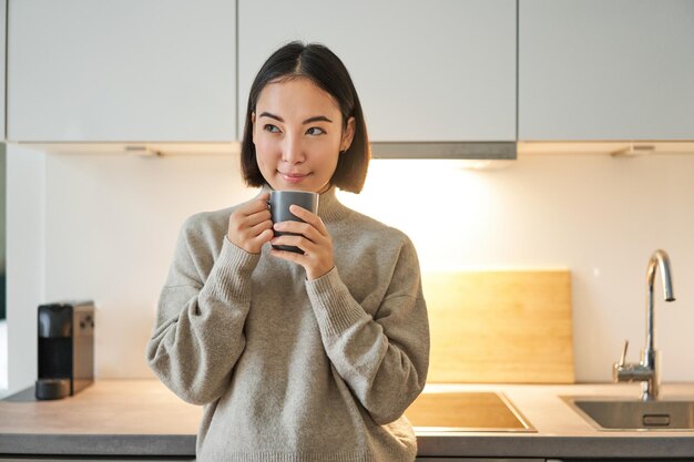 キッチンに立ってコーヒーを飲み、カメラのコンセプトを見ている笑顔のアジア人女性のポートレート