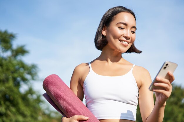 Portrait of smiling asian woman with yoga mat looking at her smartphone and reading on application s