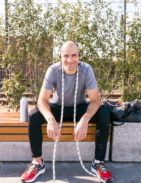 Ritratto di un uomo sorridente dell'atleta con la corda intorno al suo collo che si siede sul banco con la bottiglia di acqua e la borsa