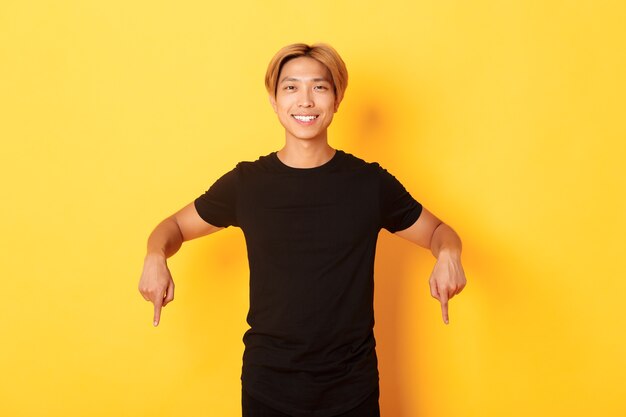 Портрет улыбающегося привлекательного корейского парня со светлыми волосами, в черном костюме, указывая пальцами вниз, показывая дорогу, стоя на желтой стене.