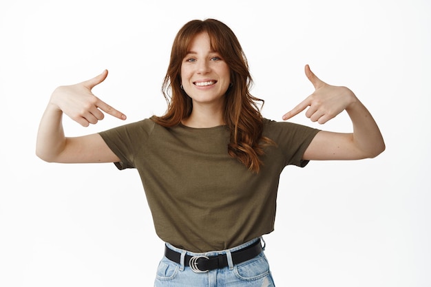 Ritratto di donna attraente sorridente che punta a se stessa, che mostra il logo sulla maglietta centrale, dimostra la pubblicità con emozione felice, in piedi su sfondo bianco