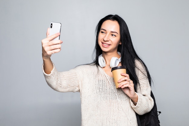 白い壁に分離されたコーヒーカップを奪うを押しながらselfieを取って笑顔の魅力的な女性の肖像画