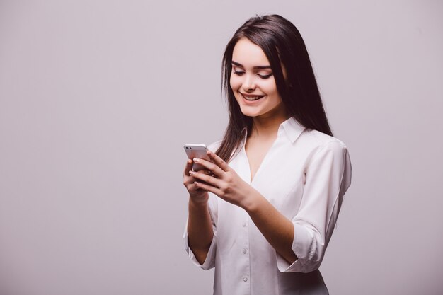 白い背景の上に孤立して立っている間携帯電話で笑顔の魅力的な女性のテキストメッセージの肖像画