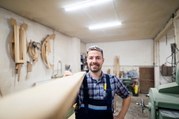 Portrait of smiling bearded carpenter woodworker holding wooden plank on shoulder ready to do his next project in carpentry workshop