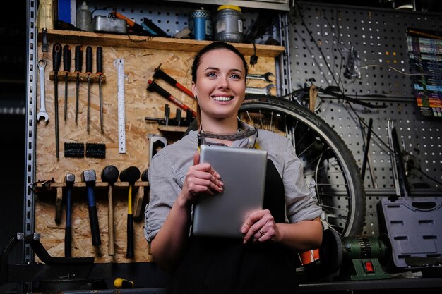 Portrait of a smiling beautiful brunette female wearing working clothes, apron and goggles, holds a tablet computer in a workshop.