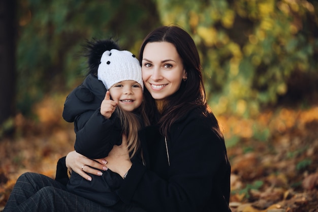 Portrait of smiling beautiful mother with her cute little daughter sitting in the autumn park. Parenthood concept