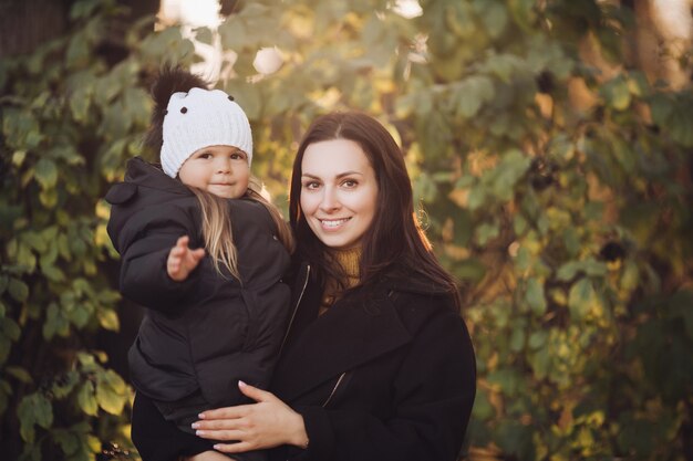 Portrait of smiling beautiful mother with her cute little daughter sitting in the autumn park. Parenthood concept