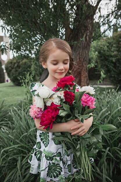 Portrait of smiling beautiful teenager girl with bouquet of peonies against green grass at summer park. Kids fashion concept.