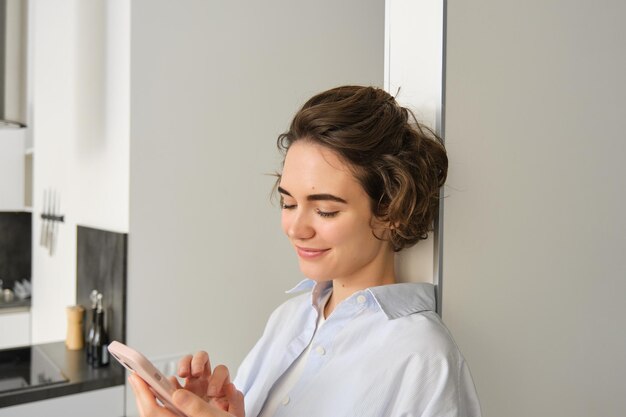 Foto gratuita ritratto di bella donna sorridente che guarda lo smartphone in piedi a casa vicino alla cucina che controlla mes