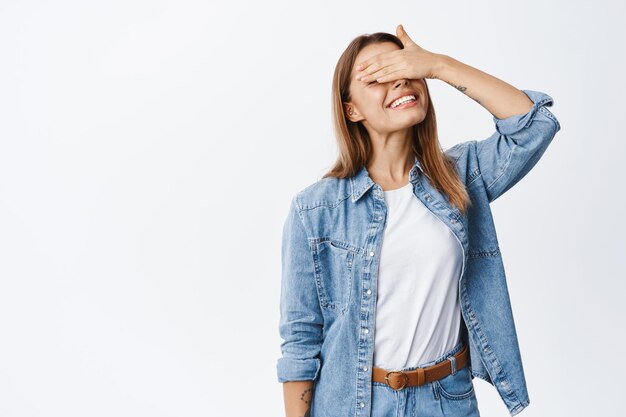 Portrait of smiling beautiful woman waiting for surprise while eyes covered with hand, anticipating something good, standing on white