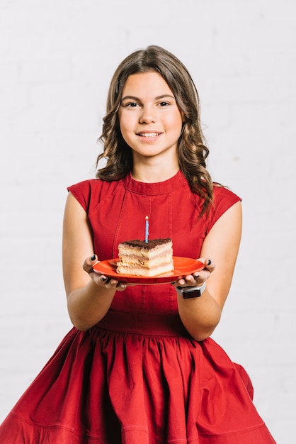 Ritratto di una ragazza sorridente di compleanno che tiene una fetta di torta sul piatto con una candela illuminata
