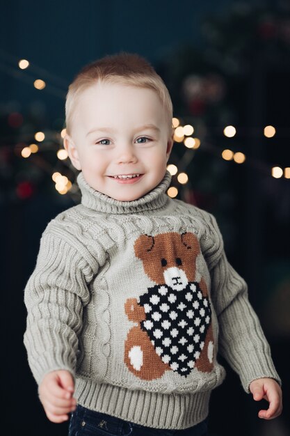 カメラを見ているテディベアと暖かいタートルネックのセーターで笑顔の金髪の赤ちゃんの肖像画。