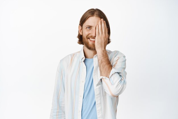 Portrait of smiling blond man cover half of face and looking happy, before after one side, standing in casual clothes against white background. Copy space