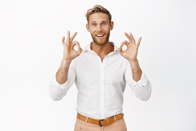 Portrait of smiling blond man in shirt showing okay signs give approval satisfied with smth standing against white background Copy space