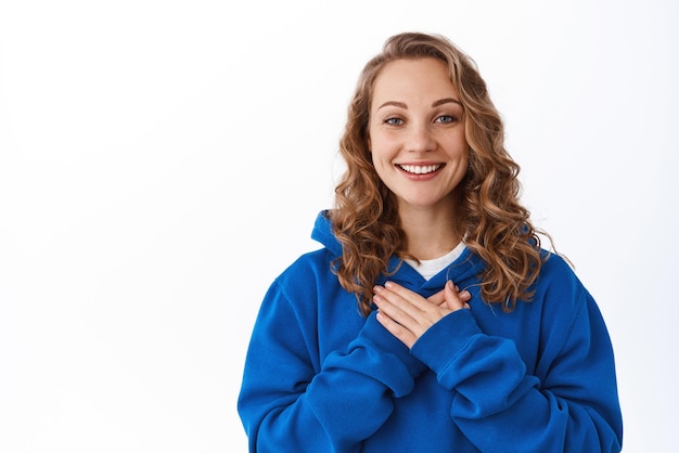 Portrait of smiling blond woman holding hands on heart and thanking you express gratitude being grateful standing over white background