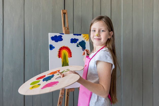 Foto gratuita ritratto di una pittura disponente sorridente della tavolozza bionda della tenuta della ragazza sul cavalletto con il pennello