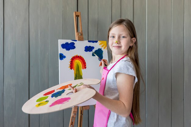 Portrait of a smiling blonde girl holding palette in hand painting on the easel with paint brush