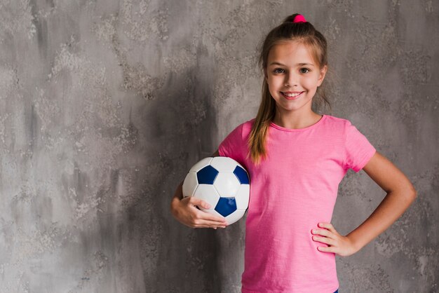 灰色の壁に対してサッカーボールを保持している腰に手で微笑んでいるブロンドの女の子の肖像画