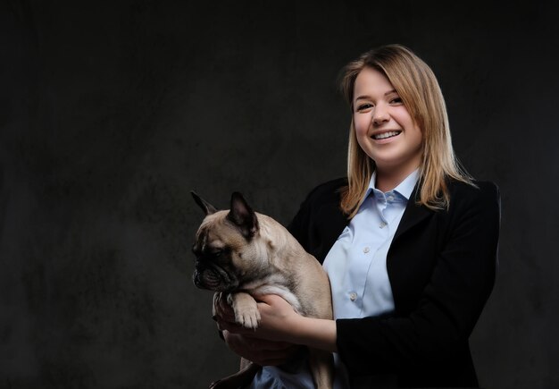 Portrait of a smiling blonde woman breeder holds her cute pug. Isolated on a dark textured background.