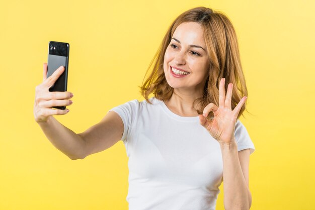 Portrait of a smiling blonde young woman taking self portrait on mobile phone making ok gesture