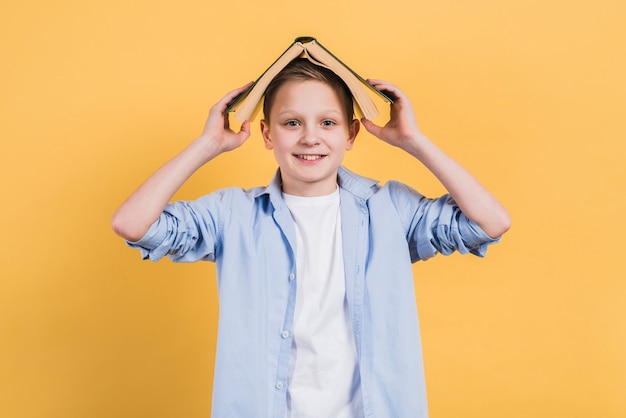 Portrait of a smiling boy holding book over his head looking to camera against yellow background