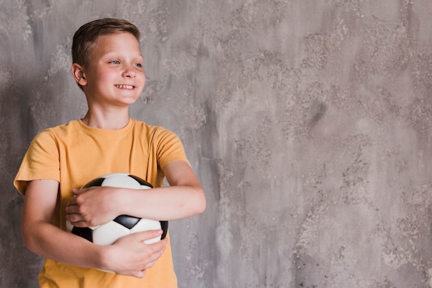 Портрет улыбающегося мальчика с футбольным мячом перед бетонной стеной